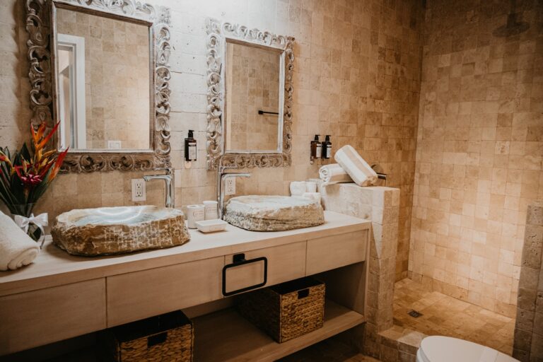 A rustic bathroom in Roatan, Honduras features stone vessel sinks set in a wooden vanity with decorative mirrors and beige tiled walls.
