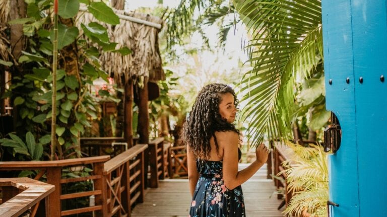 A young woman with curly hair wearing a floral dress stands on a wooden pathway surrounded by lush tropical plants in Roatan, Honduras.