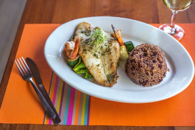 A plate of grilled fish topped with microgreens, served with rice and vegetables, is placed on an orange woven placemat next to a glass of white wine on a wooden table in Roatan, Honduras.