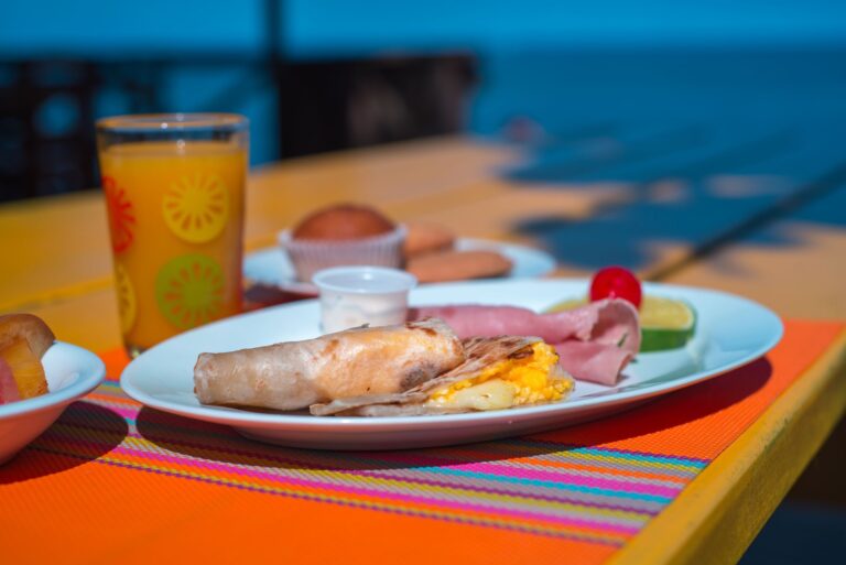 A plate with eggs, ham, and a slice of lime is set on a colorful, striped tablecloth outdoors in Roatan, Honduras, with a glass of orange juice and small bowls of condiments in the background.