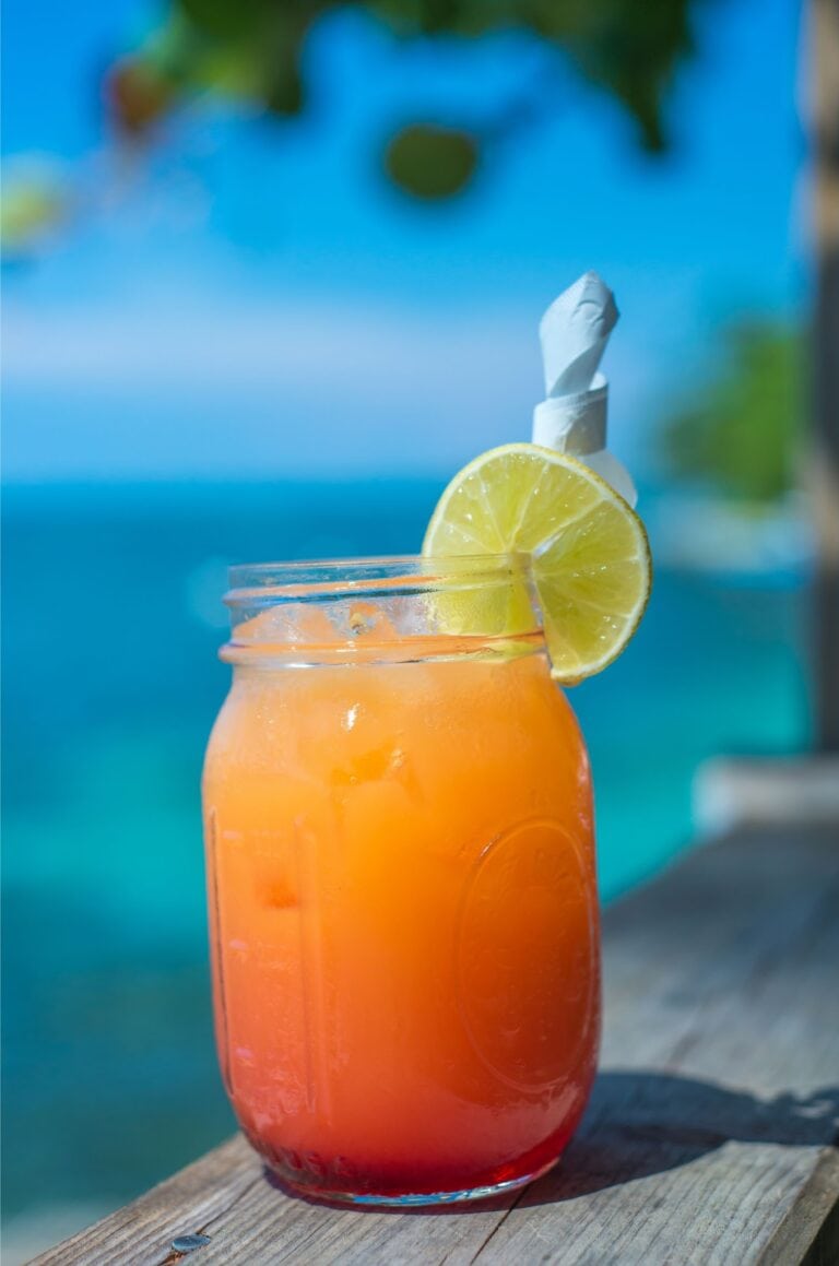 A vibrant orange and red cocktail garnished with a lemon slice sits on a weathered wooden surface against a blurred background of a swimming pool in Roatan, Honduras.