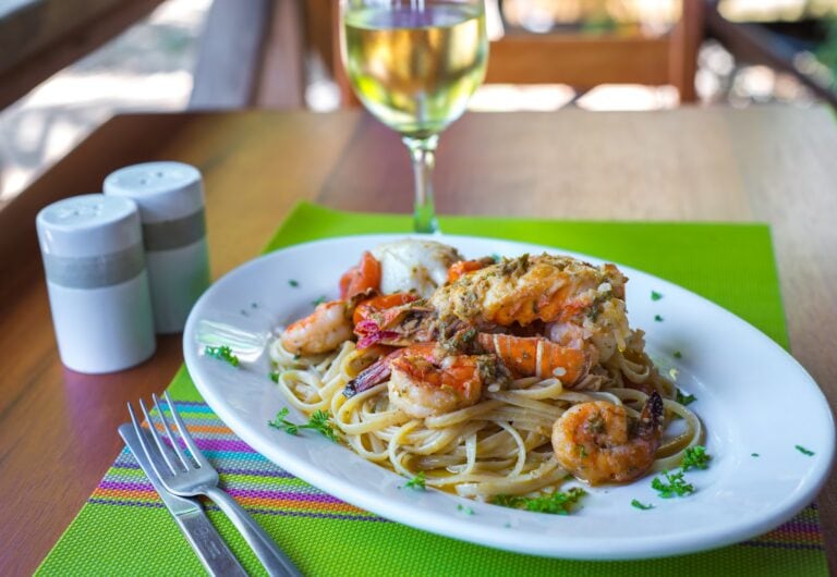 A plate of seafood pasta with shrimp and lobster sits on a colorful placemat next to a glass of white wine, with salt and pepper shakers in the background at a restaurant in Roatan, Honduras.