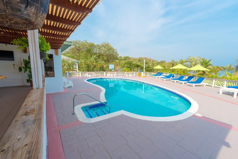 A swimming pool with a hot tub surrounds a deck area with blue lounge chairs and yellow umbrellas, overlooking lush greenery and a bright blue sky in Roatan, Honduras.