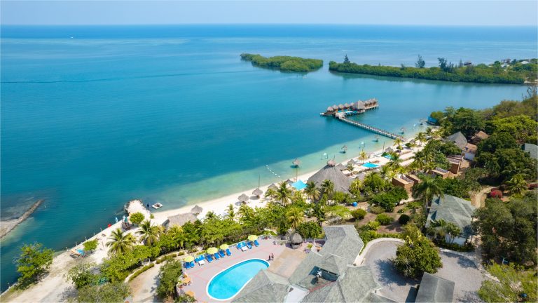 Aerial view of a tropical beach resort in Roatan, Honduras, featuring a sandy shoreline with thatched umbrellas, palm trees, a swimming pool, overwater bavilions, and lush greenery overlooking the Caribbean Sea.