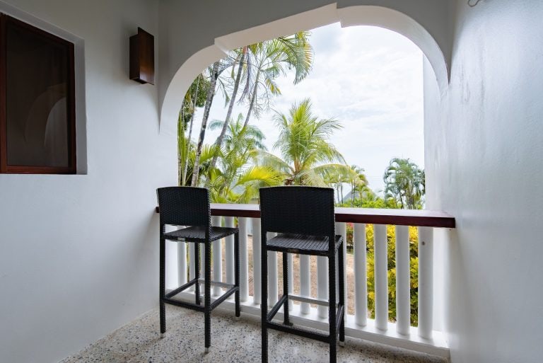 A balcony with three black woven chairs overlooking lush green palm trees on Roatan, Honduras, with a white railing and an arched opening framing a tropical view.