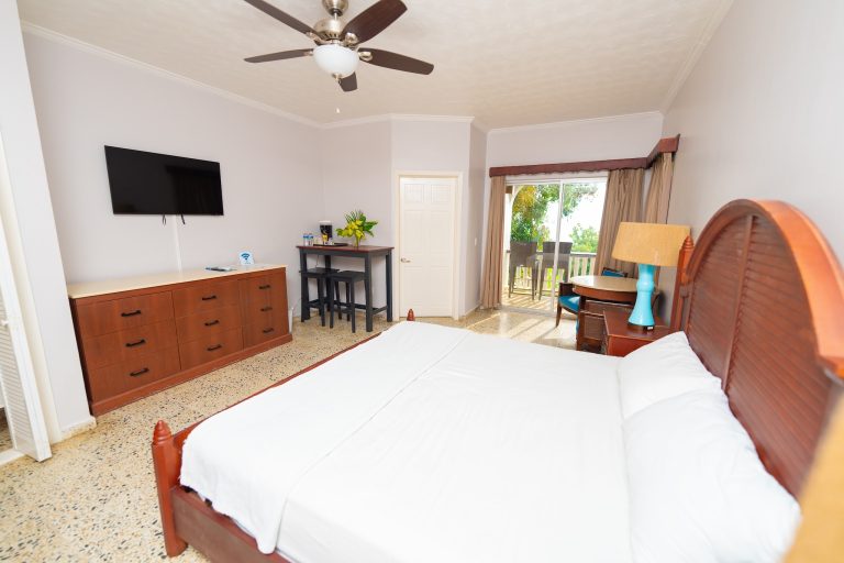 A bedroom in a hotel or vacation rental on Roatan, Honduras features a wooden bed frame with white bedding, a flat-screen TV mounted on the wall, a small desk with water bottles and a plant, a sliding glass door leading to a balcony with outdoor seating and greenery, and neutral-colored decor with natural light.