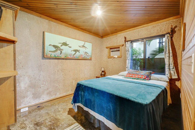 A cozy bedroom in Roatan, Honduras, features a bed with colorful pillows, a window with tropical plants outside, wooden-paneled walls and ceiling, and a framed underwater sea turtle artwork on the textured wall.