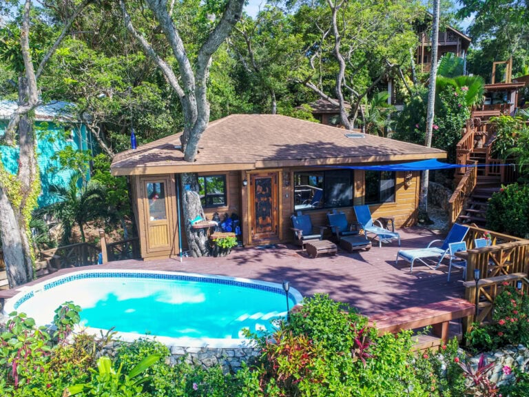 A cozy wooden cottage with a brown shingle roof is surrounded by lush tropical trees and plants on Roatan in Honduras, featuring a private deck with lounge chairs near a small swimming pool.