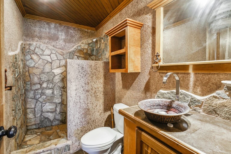 A rustic bathroom in Roatan, Honduras features a stone shower area with natural rock walls, a wooden ceiling, a vessel sink with a patterned bowl, a wooden cabinet and mirror frame, and textured plaster walls.