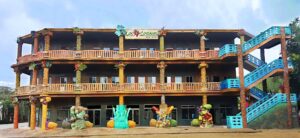 The colorful, multi-story hotel in Roatan, Honduras, features vibrant tropical decorations, wooden balconies, and bright blue staircases, with a sign that reads "Los Corales" at the top.