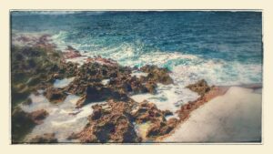 Rough volcanic rocks along the shoreline with ocean waves crashing against them on a sunny day in Roatan, Honduras.