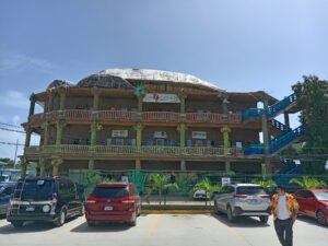 A multilevel hotel with a coral reef and turtle-themed decor located in Roatan, Honduras, overlooking a parking lot filled with cars and a person walking by under a clear blue sky.