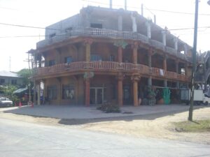 A building under construction in Roatan, Honduras, features colorful wooden balconies and decorative elements on its exterior, with utility poles and parked vehicles visible nearby.