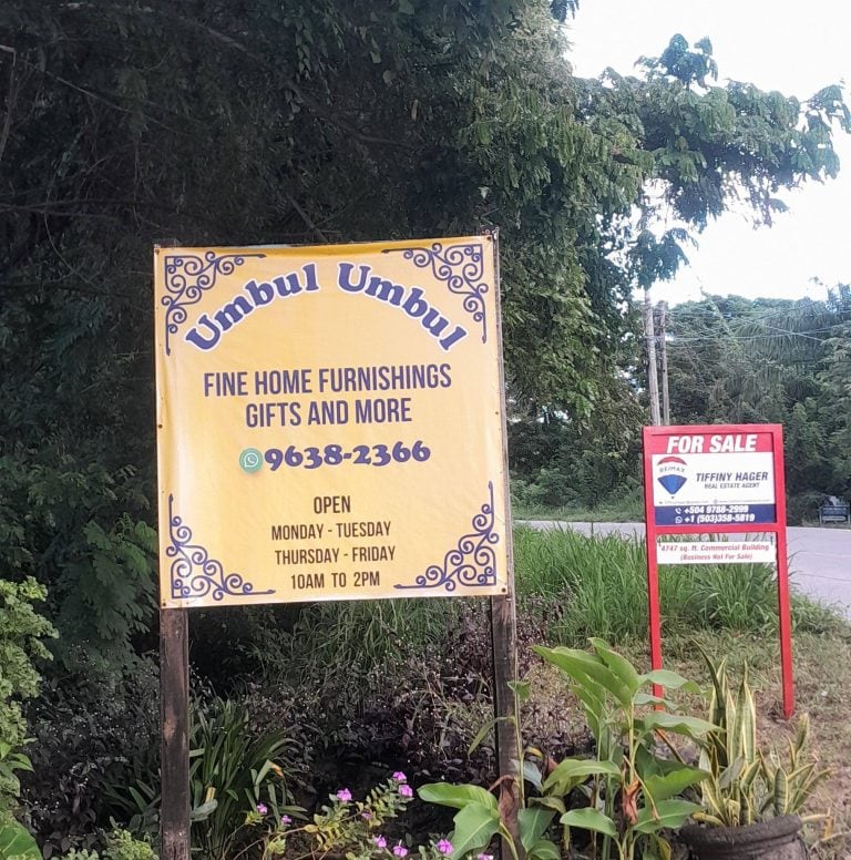 A yellow sign advertising "Umbul Umbul" fine home furnishings and gifts is displayed among lush green foliage on Roatan, Honduras, with additional signs for real estate and sales nearby.