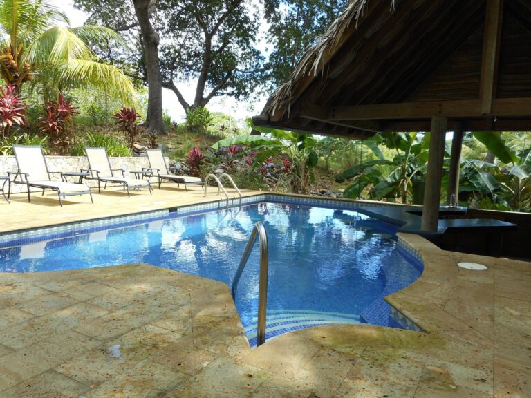 A swimming pool area surrounded by tropical plants and lounge chairs, with a thatched-roof gazebo and lush greenery in Roatan, Honduras.