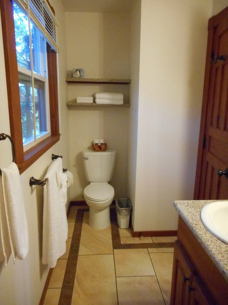 A small bathroom in a house on Roatan, Honduras, features a toilet, two wall-mounted shelves with folded towels, a window with wooden trim, and a granite countertop with a sink, with towels hanging on a rod next to the window.