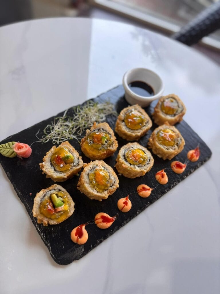 Sushi rolls with avocado and sauce garnishes are arranged on a black slate serving platter, accompanied by a small dish of soy sauce and decorative edible flowers and sprouts, set on a white table in Roatan, Honduras.