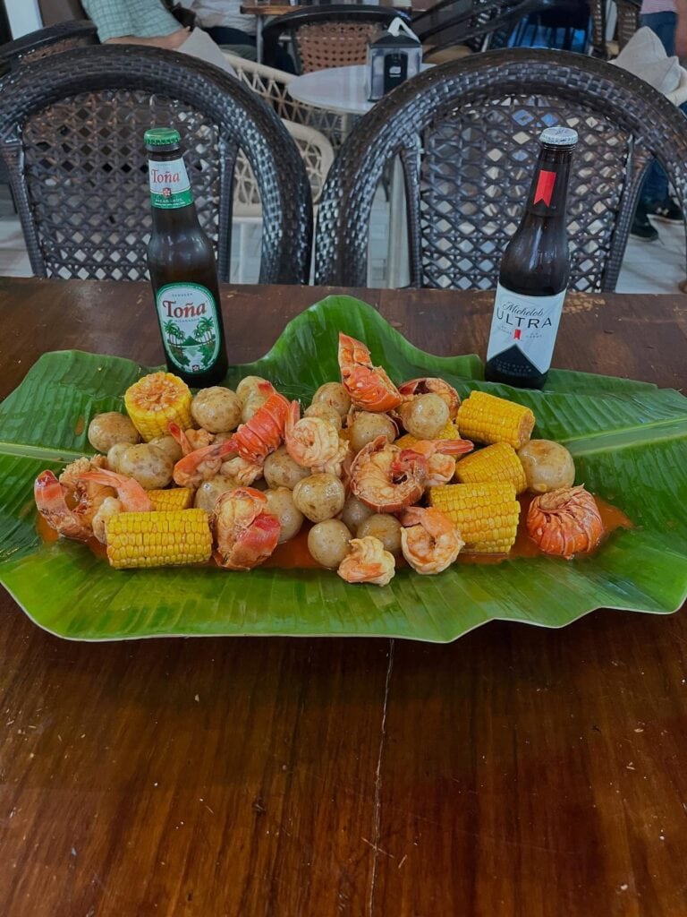 A large green banana leaf serves as a platter for a seafood dish featuring boiled shrimp, corn on the cob, and small potatoes, with two bottles of Honduran beer placed on a wooden table in a casual restaurant setting in Roatan, Honduras.