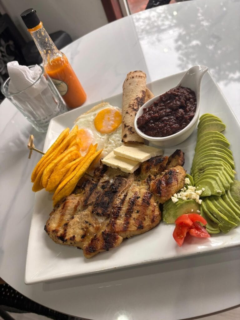 A plate of Honduran cuisine featuring grilled chicken, fried plantains, a fried egg, refried black beans, sliced avocado with cheese crumbles, cherry tomatoes, a rolled tortilla, and a side of hot sauce, served on a white dish on a white table in Roatan.
