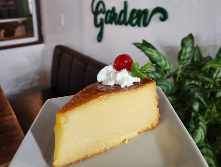 A slice of creamy cheesecake topped with whipped cream, a cherry, and a small mint leaf is displayed on a white plate in a restaurant setting on Roatan, Honduras.