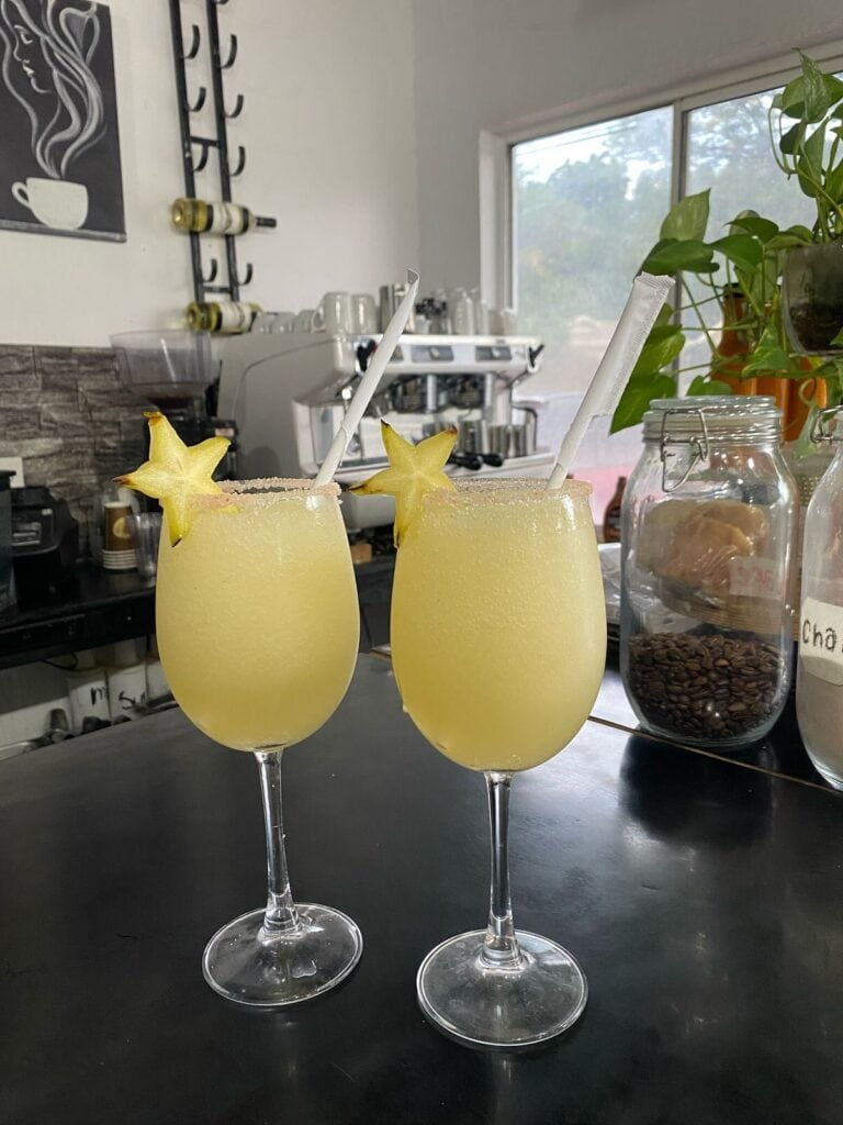 Two yellow tropical cocktails with starfruit garnishes and straws are displayed on a black counter inside a cafe or bar in Roatan, Honduras, with a coffee machine, jars, and a plant visible in the background.