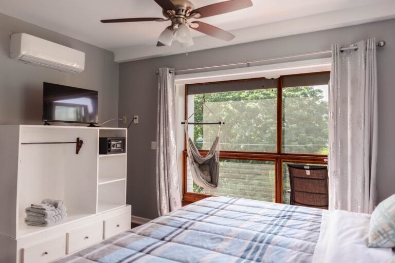 A bright, airy bedroom in Roatan, Honduras features a large window with white curtains, a swinging hammock chair, a ceiling fan, a wall-mounted air conditioner, and a white shelving unit with neatly folded towels, all with a view of lush greenery outside.