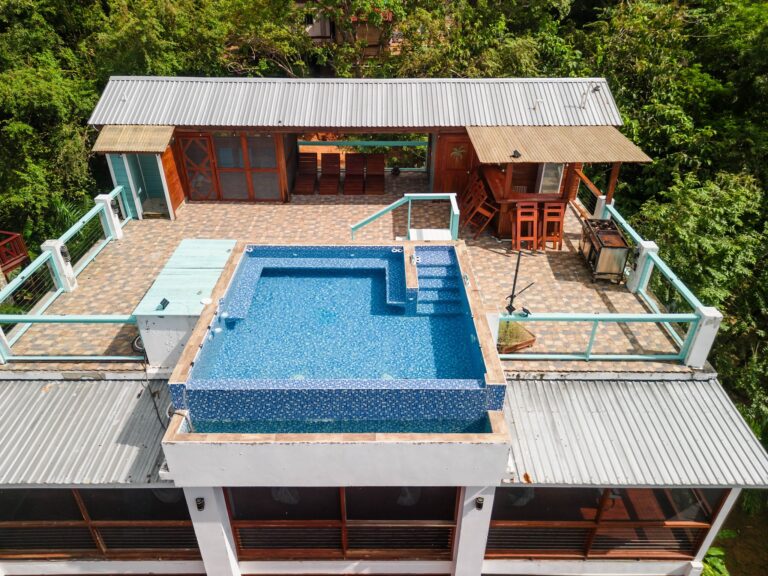 A rooftop terrace in Roatan, Honduras, features a swimming pool, wooden seating area, barstools, and is surrounded by lush green trees.
