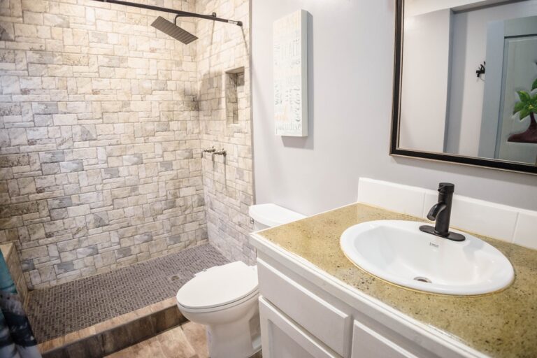 A compact bathroom in Roatan, Honduras, features a stone-tiled walk-in shower with a rainfall showerhead, a white toilet, a beige countertop with a white sink and black faucet, a large mirror, and decorative wall art.