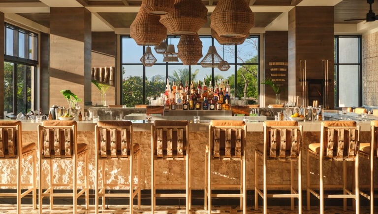 A modern indoor bar in Roatan, Honduras, features a white marble counter with wooden barstools, a central display of assorted liquor bottles, large woven pendant lights hanging from the ceiling, and large windows showcasing lush greenery outside.