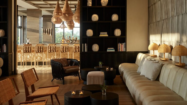 A stylish interior lounge area in Roatan, Honduras, featuring a cream-colored sofa with decorative pillows, wooden tables with candles, rattan lamps along the wall, a black bookshelf with decorative vases and books, and a view of a bar area with high chairs and hanging wicker pendant lights.
