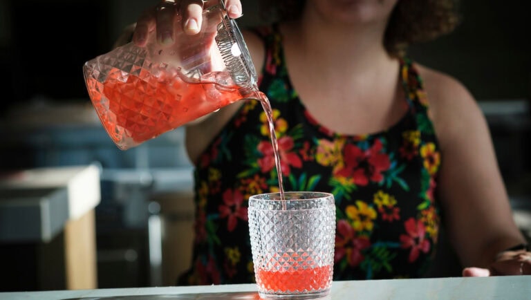 A person in a colorful floral top is pouring a pink cocktail from a textured glass pitcher into a matching textured glass on a bar counter in Roatan, Honduras.