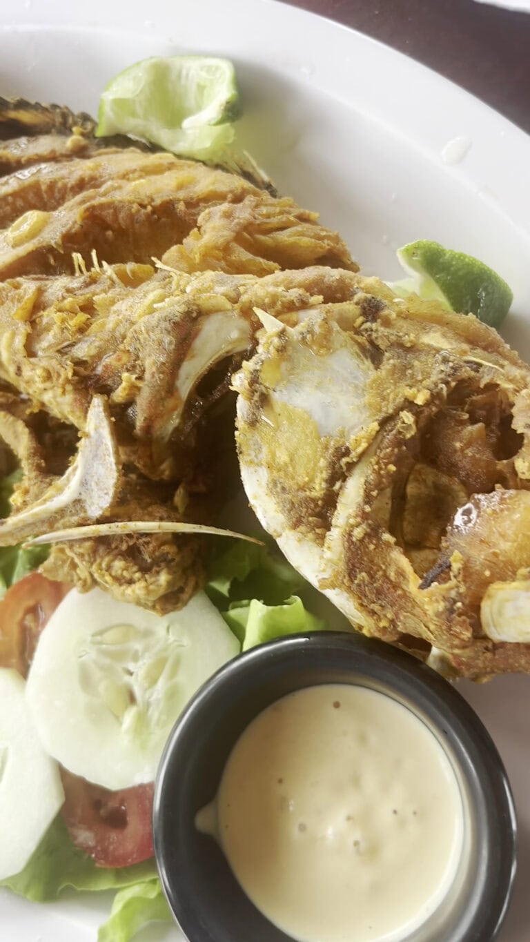 Fried fish served with lime wedges, sliced tomatoes, lettuce, and a cup of creamy dipping sauce on a white plate, typical of Roatan, Honduras.