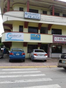 A building in Roatan, Honduras, with signs for businesses including a foundation, an optical shop, and a barber shop, parked cars in front, and a pedestrian crosswalk on the street.