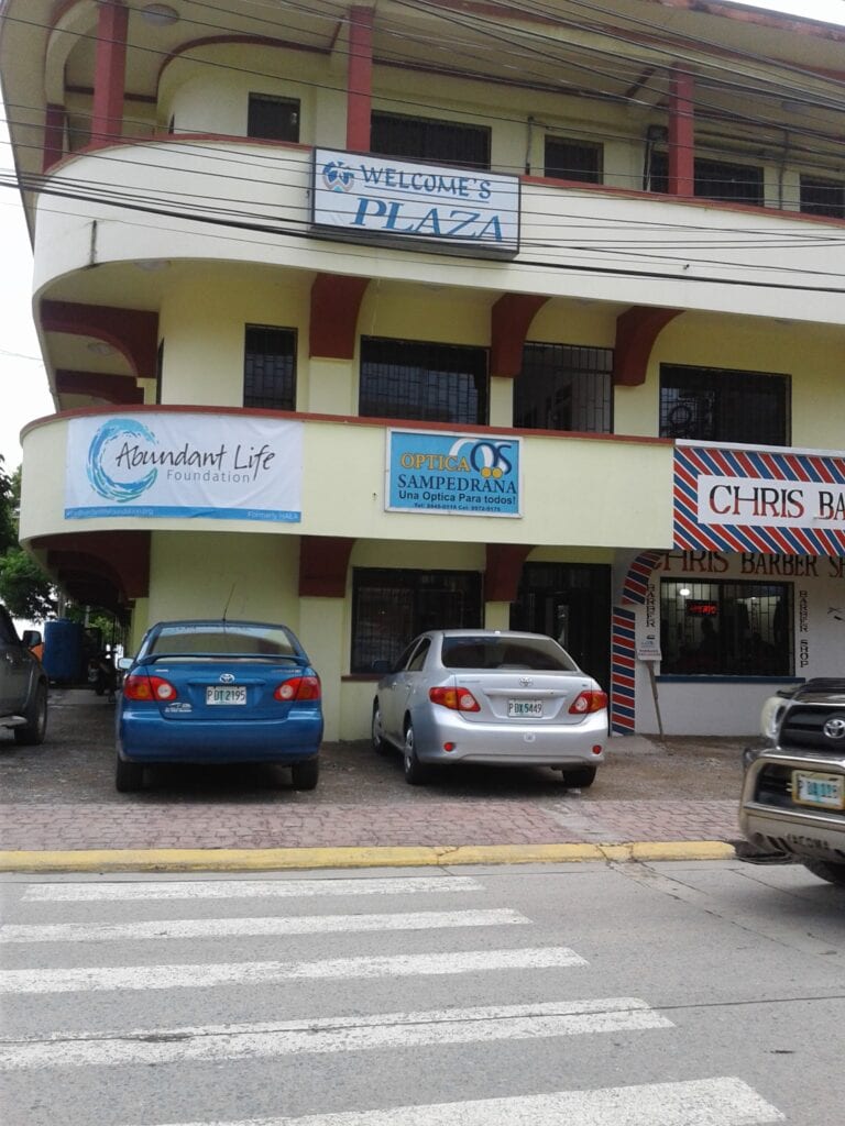 A building in Roatan, Honduras, with signs for businesses including a foundation, an optical shop, and a barber shop, parked cars in front, and a pedestrian crosswalk on the street.