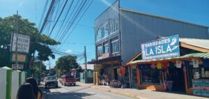A busy street scene in Roatan, Honduras, featuring a colorful storefront with a sign for "Variedades La Isla," bicycle rentals, and overhead power lines against a bright blue sky.