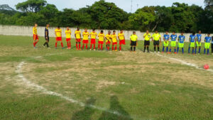 A soccer match with players in yellow and red uniforms and blue and neon yellow uniforms taking place on a grassy field in Roatan, Honduras, surrounded by a concrete wall and lush green trees.