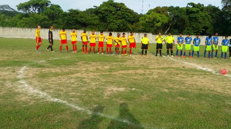 A soccer match with players in yellow and red uniforms and blue and neon yellow uniforms taking place on a grassy field in Roatan, Honduras, surrounded by a concrete wall and lush green trees.