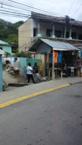 A small outdoor market stall on a street in Roatan, Honduras, selling colorful clothing and accessories, with people browsing and interacting near a residential building and laundry hanging on a fence.