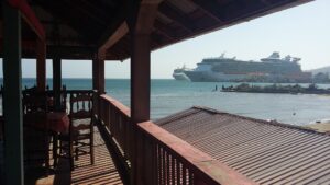 A view from a wooden seaside restaurant or bar in Roatan, Honduras, showing two large cruise ships anchored in the Caribbean Sea with a small building and palm trees on the shore in the background.