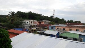 A view of a small town in Roatan, Honduras, featuring colorful buildings, metal roofs, lush green hills, and a communication tower under a cloudy sky.