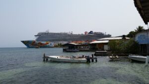A large cruise ship with colorful artwork on its hull is docked near small boats and waterfront buildings in Roatan, Honduras.