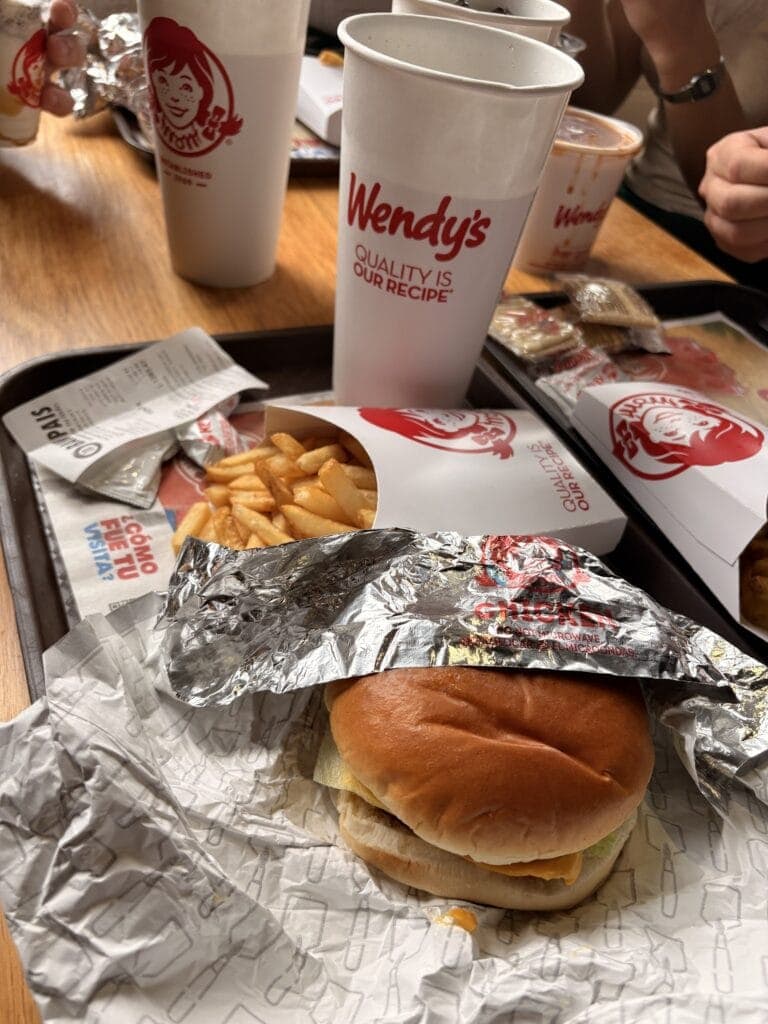 A Wendy's fast food meal showing a cheeseburger wrapped in foil, a side of French fries, and three large drinks on a wooden table in Roatan, Honduras.