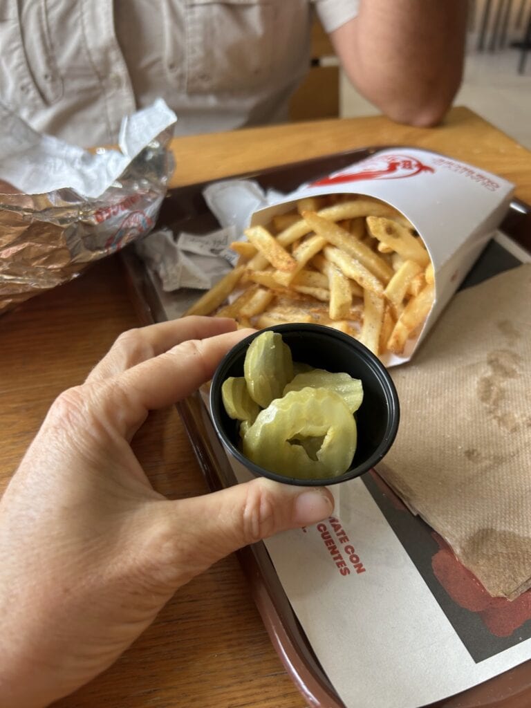 A person holds a cup of pickled green vegetables next to a serving of French fries in a white paper tray at a casual dining establishment in Roatan, Honduras.