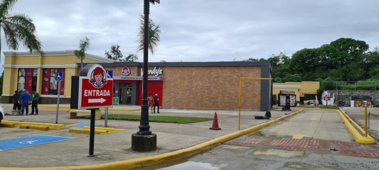 A Wendy's fast food restaurant with a yellow and gray exterior is situated in Roatan, Honduras, featuring a red and white sign, a wheelchair-accessible parking area, and a small group of people near the entrance, with lush green trees in the background.