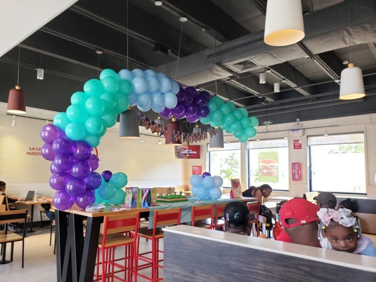 Children and adults sit at tables decorated with colorful balloons and birthday banners inside a restaurant or event space in Roatan, Honduras, with large windows and modern ceiling lights.