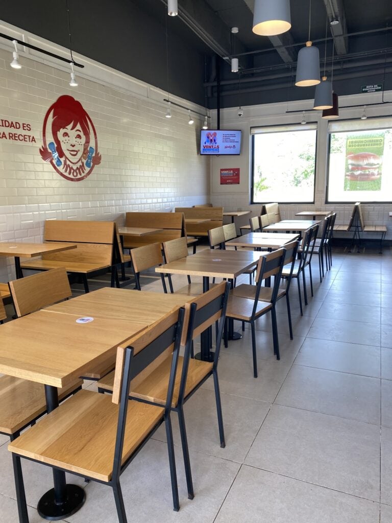 The image shows the interior of a modern restaurant or café in Roatan, Honduras, with wooden tables and chairs arranged near large windows, white brick walls, and contemporary pendant lighting.