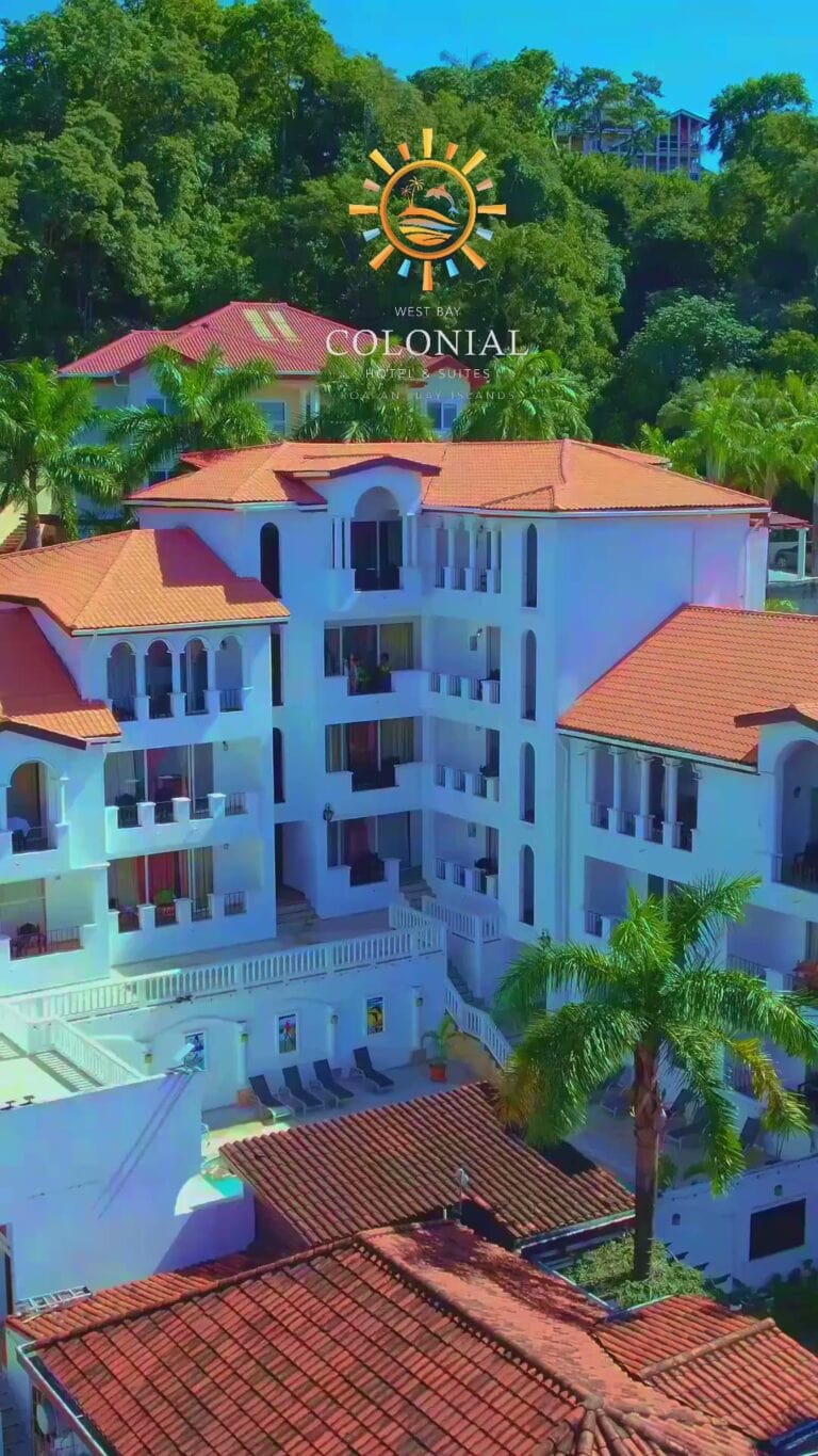 An aerial view of the Colonial Hotel & Suites in Roatan, Honduras, featuring a white multi-story building with red-tiled roofs and balconies surrounded by lush green palm trees and tropical foliage.