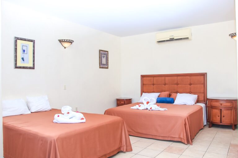 A hotel room in Roatan, Honduras, features two double beds with orange bedspreads, decorative towel animals shaped like swans, and wall-mounted light fixtures, with white walls decorated with framed artwork and an air conditioning unit.