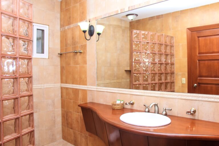 A bathroom in Roatan, Honduras featuring beige tiled walls, a large mirror, a glass block partition, a wooden vanity with a white sink, and a small window with frosted glass.