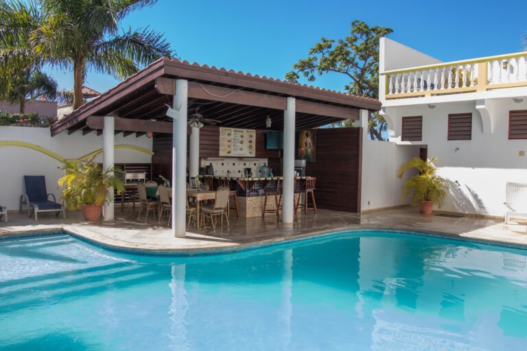 A swimming pool area with a covered outdoor bar and seating area featuring a menu, surrounded by tropical plants and white buildings with balconies in Roatan, Honduras.